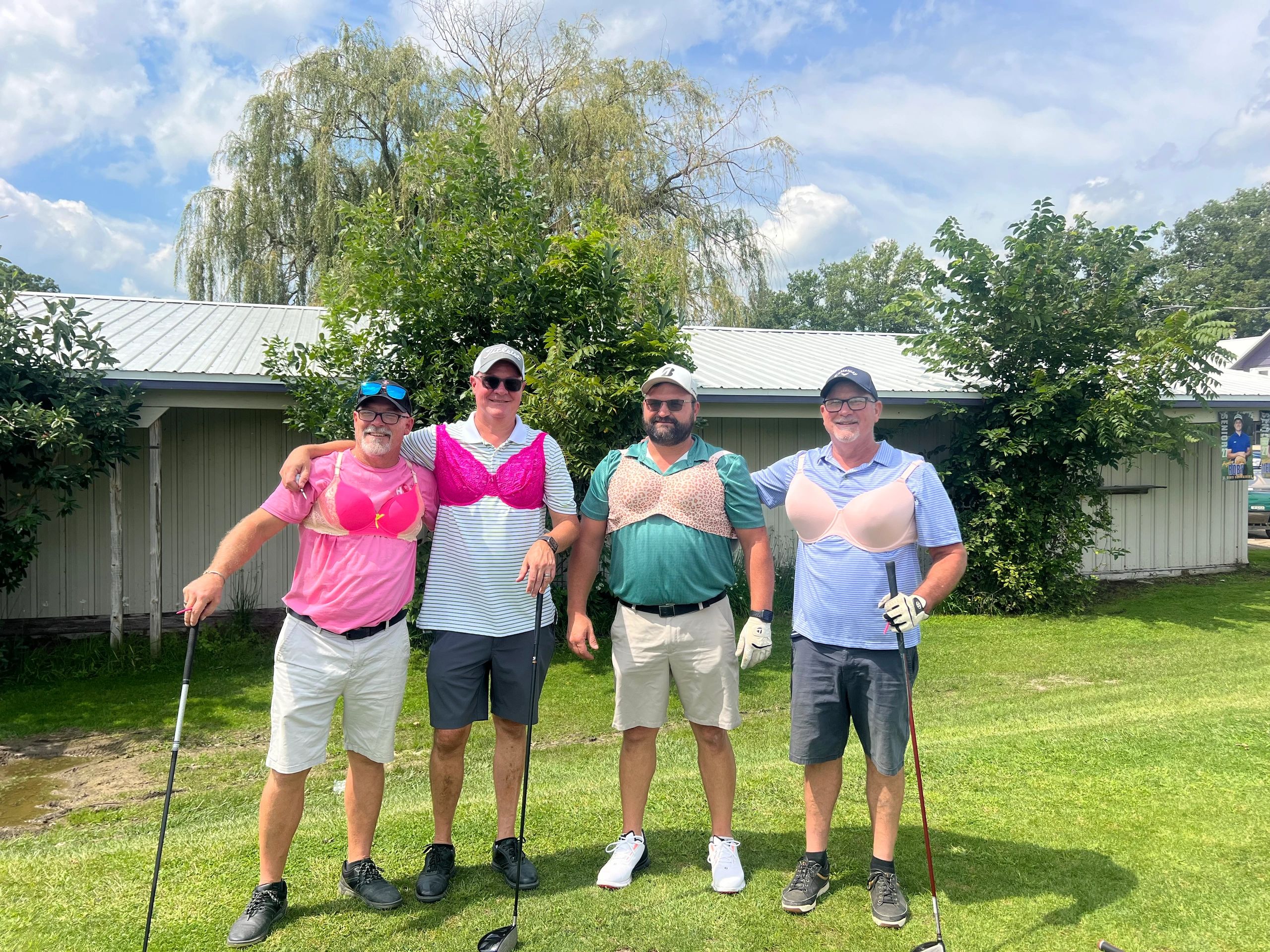 Four men posing with bras over their golf clothes on a sunny day.