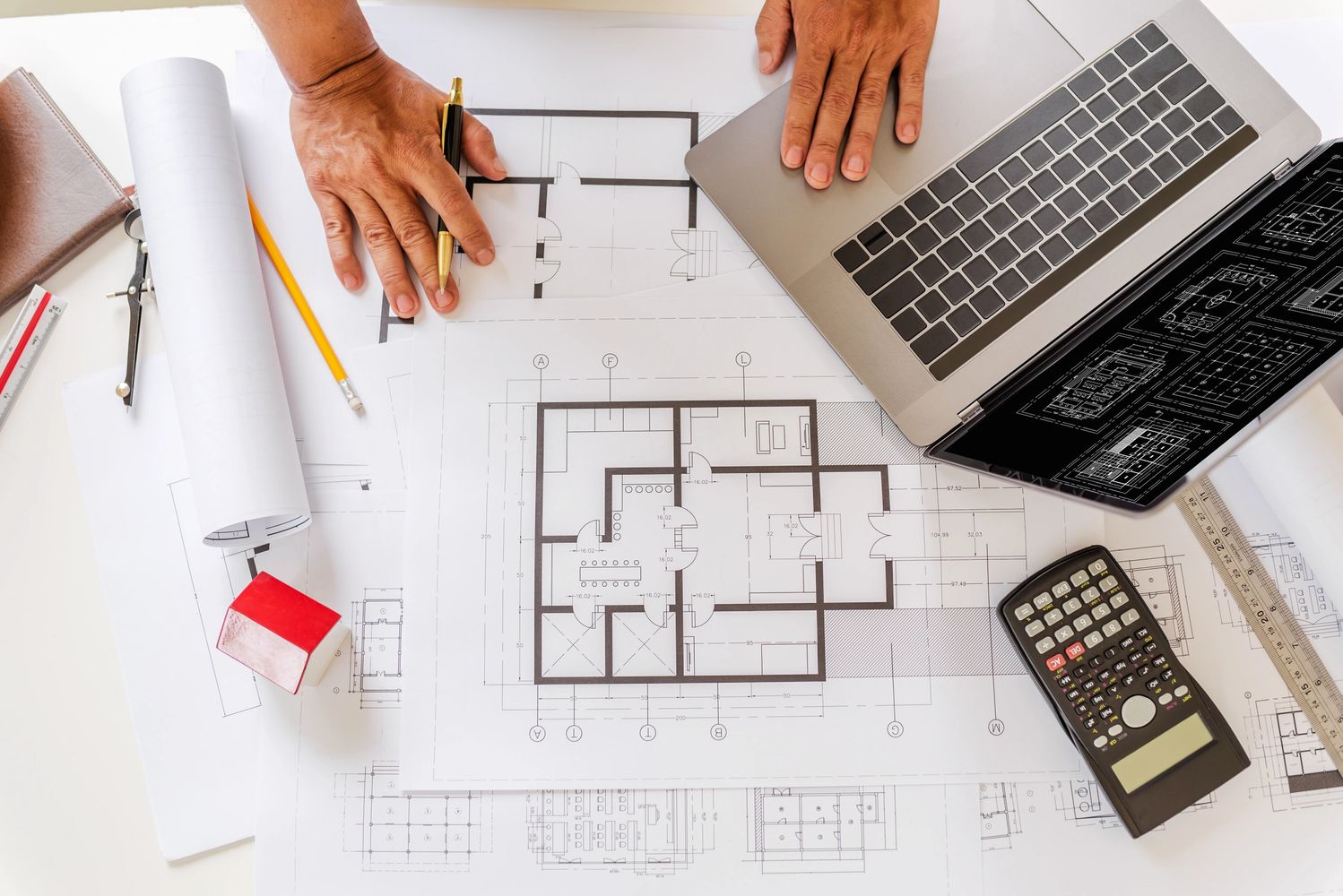 Hands working on architectural blueprints with a laptop, calculator, and drafting tools.