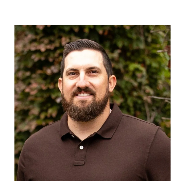 Man with beard smiling in an outdoor setting with greenery behind him.