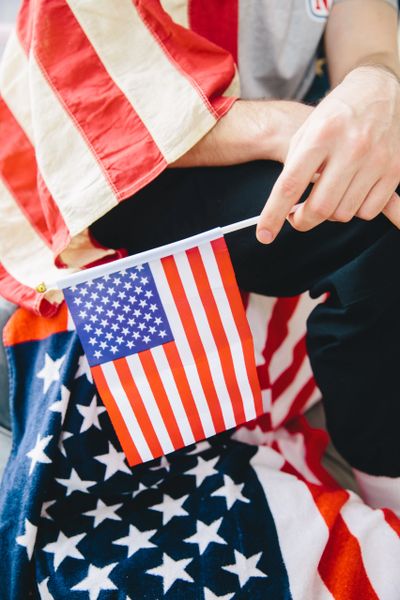Someone sits down and holds a small American flag. They are blanketed in a larger American flag. 