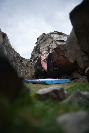 Man bouldering shirtless on a rocky cliff with a crash pad below.