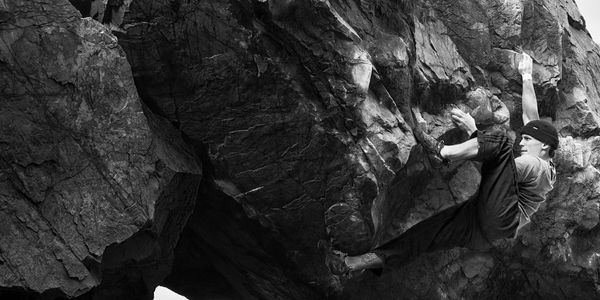 A man bouldering on a steep rock face wearing a beanie and chalked hands.