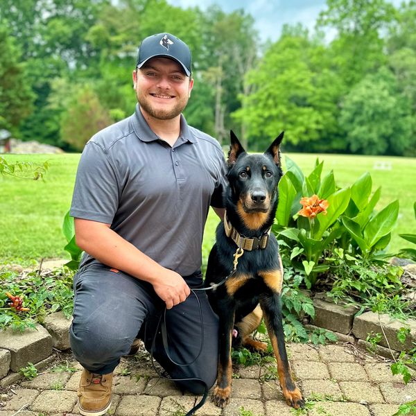 Man kneeling outdoors with a Doberman dog on a leash.