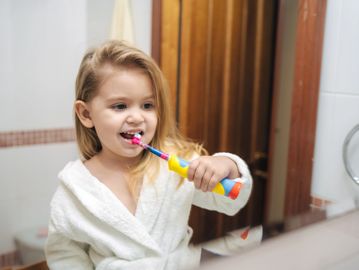 You girl in bathroom smiles as she brushes teem with with an electric toothbrush.