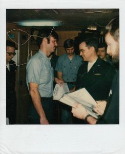 Men in casual and military attire interacting indoors, holding papers.
