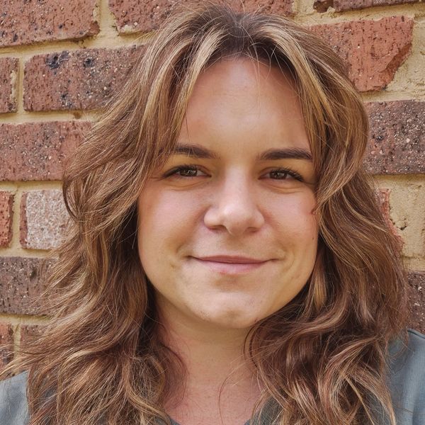 Rhiannon Pigdon, Physiotherapist at Forever Strong Physiotherapy. Smiling woman with wavy hair stands in front of a brick wall.