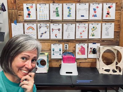 Woman smiling in front of a creative workspace with colorful artwork displayed.