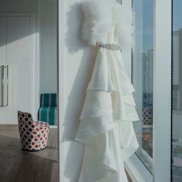 Elegant white wedding dress with a feathered shawl hangs by a bright window.