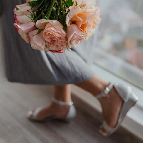 Person holding a bouquet of pink roses wearing silver sandals and gray pants.