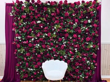A white chair in front of a lush floral wall with deep red and white flowers.