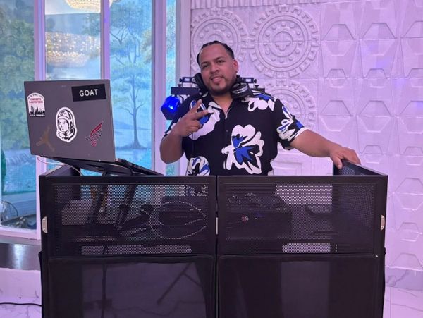 DJ posing behind his equipment with a peace sign in a stylish floral shirt.