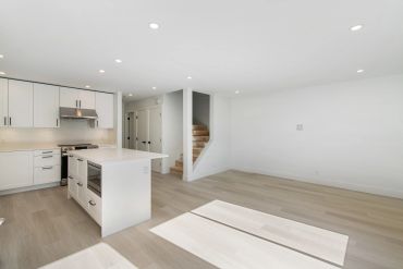 Bright modern kitchen with white cabinets and island opens to empty living space.