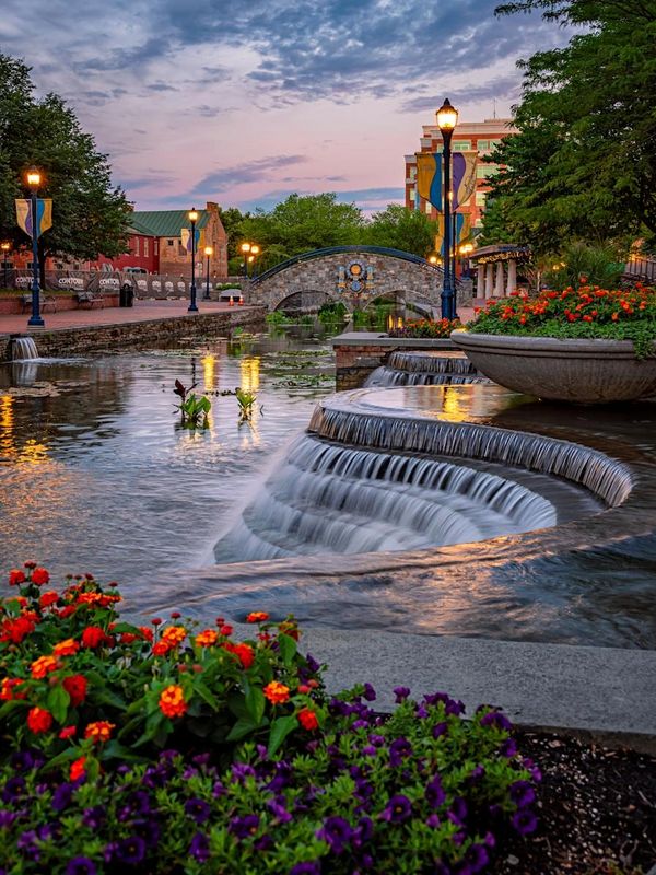 A serene urban park with cascading water, colorful flowers, and lamp-lit pathway at sunset.