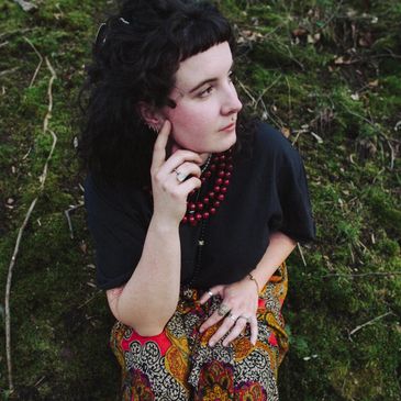 A thoughtful woman sitting on grass, wearing colorful pants and black top.