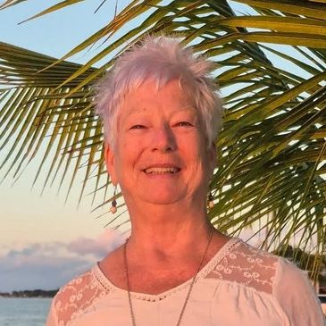 Smiling older woman outdoors with palm leaves in background.