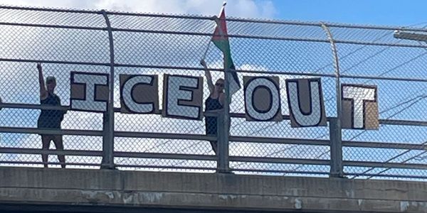 Protesters holding signs reading 'ICE OUT' on a bridge.