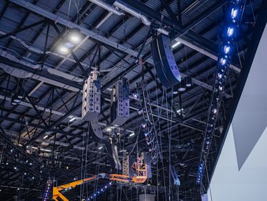 Technicians install speakers and lighting at a large event venue.