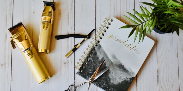 Gold hair clippers, scissors, and a notebook on a wooden surface.