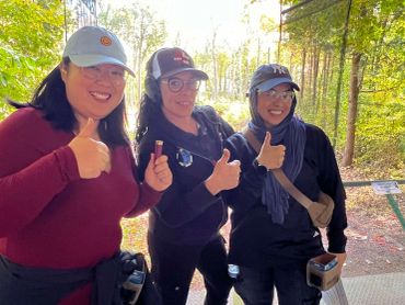 Three women outdoors giving thumbs up, smiling happily.