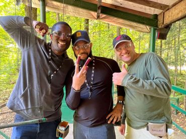 Three men posing and smiling outdoors under a wooden shelter.