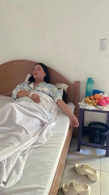 A woman resting in bed with fruit and medicine nearby on a bedside table.