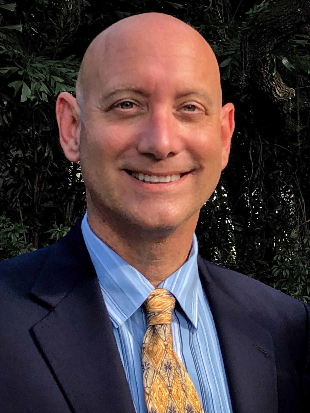 Smiling bald man in suit with yellow patterned tie outdoors.