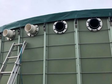A green industrial tank with multiple pipe openings and a ladder leaning against it.