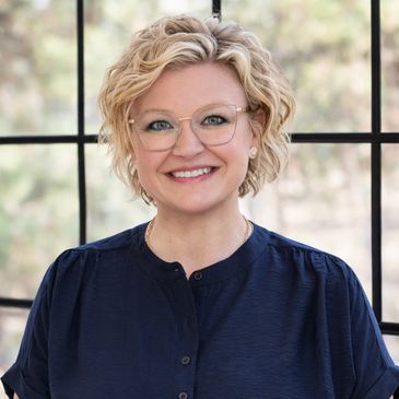 Smiling woman with glasses and curly blonde hair, wearing a navy blouse.