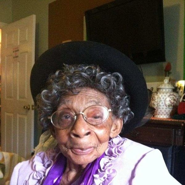 Elderly woman with curly hair, glasses, and a black hat, dressed in a lavender outfit.