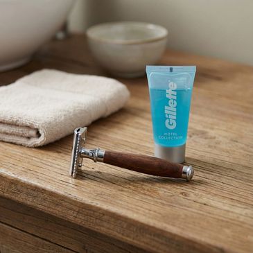 A wooden-handled razor and Gillette shaving gel on a wooden surface with towel and bowl.