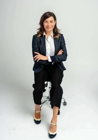 Confident woman in business attire sitting on a stool, arms crossed, smiling.