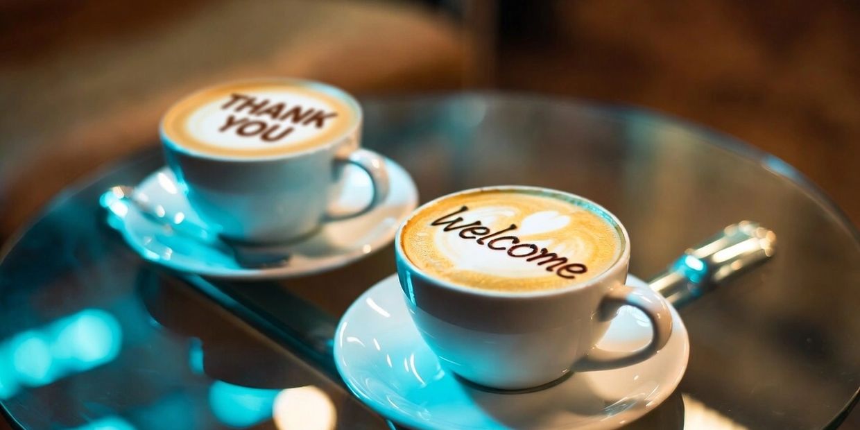 Two coffee cups with 'Thank You' and 'Welcome' written in the foam on a glass table.