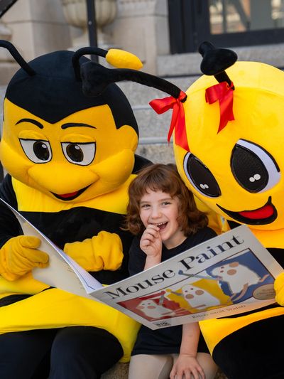 Child with two bee mascots reading a book together.