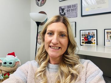 Smiling woman with curly blonde hair in a cozy room with framed photos and a lamp.