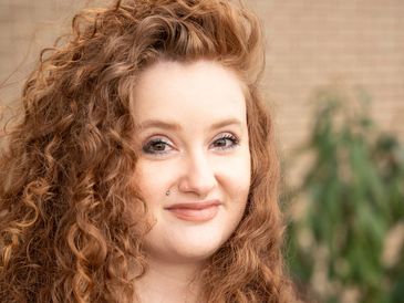 Portrait of a smiling woman with curly red hair and nose piercing.
