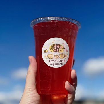 Hand holding a red drink with a 'Little Cups Big Hearts' label against a blue sky.