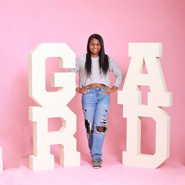Young woman posing with large graduation letters on pink background.