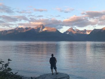 A man stands on a rock by a lake, gazing at sunlit mountains.