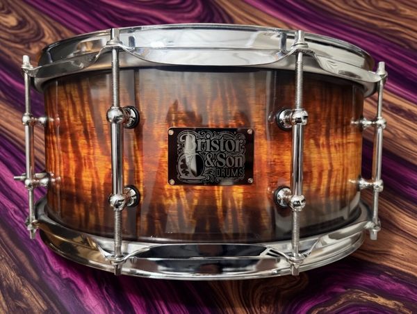 A polished Bristor &amp; Son wooden snare drum on a vibrant purple and brown surface.