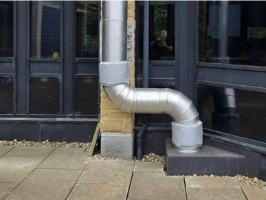 Metallic vent pipe bends sharply outside a building with black window frames.