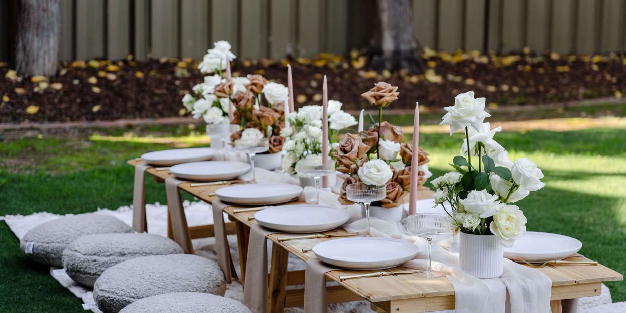 Elegant outdoor table setting with white and brown roses and soft cushions.
