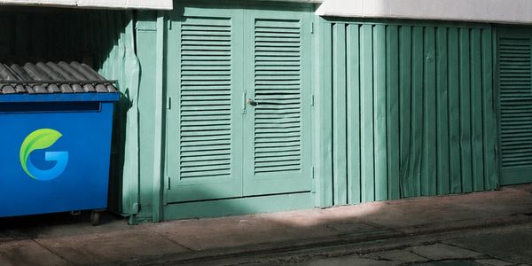 Blue dumpster with green logo next to green shutter doors on a street.