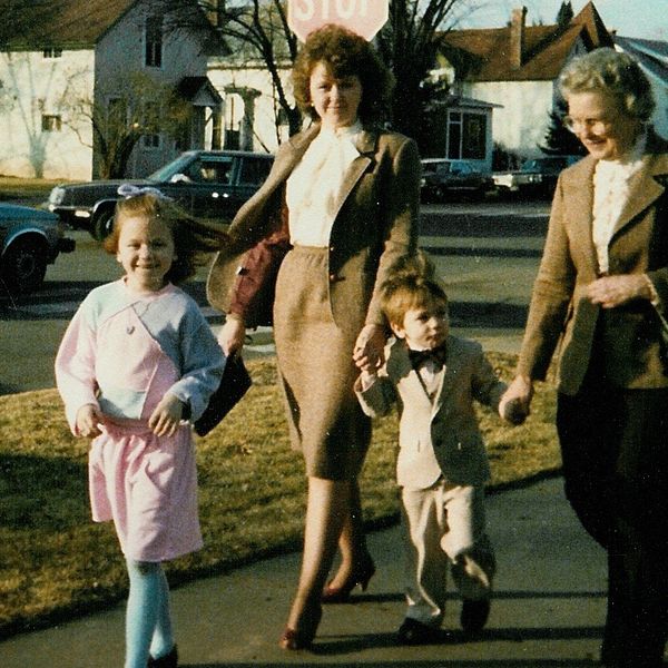 Smiling girl in pink and blue struts in front of her tan-clad mom, brother, and grandmother.
