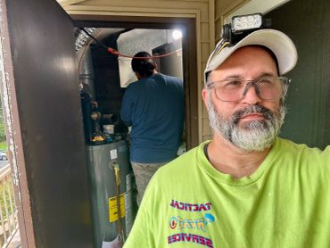 Man taking a selfie near a water heater with a worker in background.