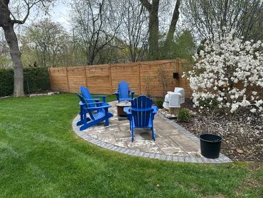 Blue Adirondack chairs arranged around a fire pit on a stone patio in a backyard.