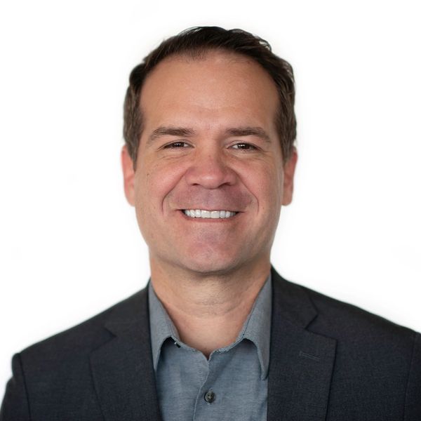 Smiling man in a dark blazer and gray shirt against a white background.