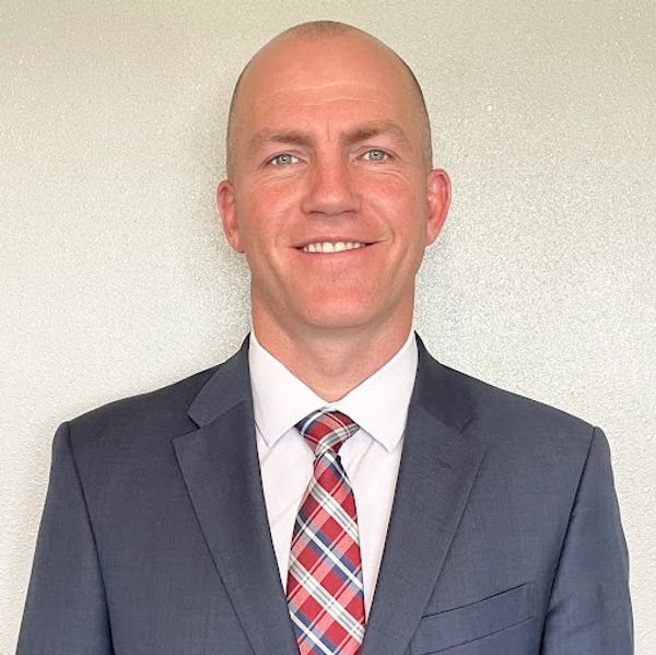 Smiling man in a business suit and patterned tie against a plain background.
