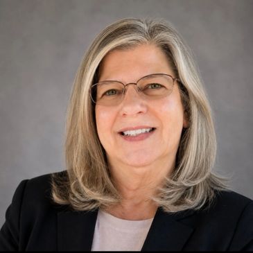 Portrait of a smiling woman with glasses and gray hair in business attire.