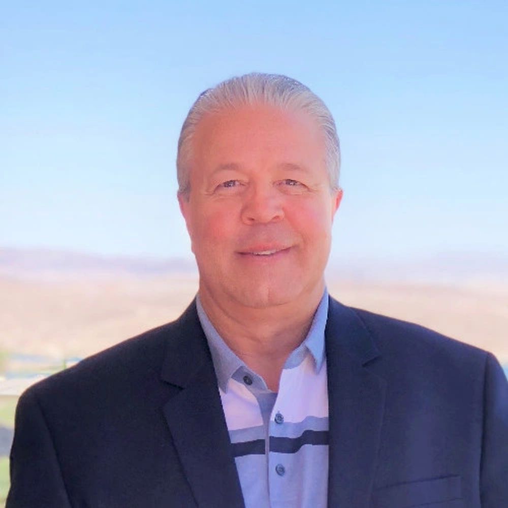 A man in a suit jacket smiling with a blurred outdoor background.