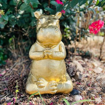Golden hippopotamus statue sitting peacefully outdoors among pine cones and flowers.
